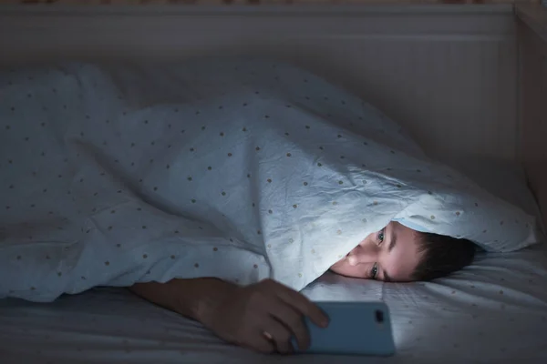 Uykulu yorgun bir adamın portresi battaniyenin altında uzanıyor ve gece yarısı akıllı telefon kullanıyor, uyuyamıyor. Uykusuzluk, nomofobi, uyku bozukluğu konsepti. Bir cep telefonuna bağımlılık. Yumuşak odak.
