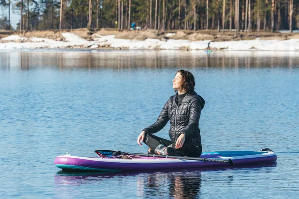 Sakin güzel kadın gölün ortasında Sup tahtasında oturuyor. Dişi yaslanarak ayakta duran kürek tahtası üzerinde. Bir çeşit meditasyon ve rahatlama. Kış mevsimi ve aktif eğlence konsepti.