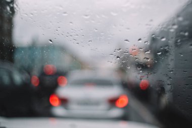 Ön cama düşen yağmur damlalarının yakın fotoğrafını çek. Seçici odak, arka planda trafik sıkışıklığında bulanık arabalar. İnce cam ısıtma iplikleri. Yağmur mevsimi. Otomotiv endüstrisi. Arabanın içindeki samimiyet..