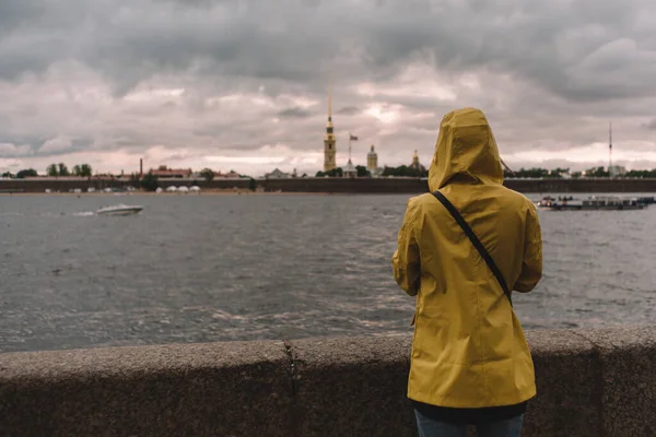 Sarı yağmurluklu bir kızın arkasından bak. Gezgin, Neva nehrine ve Saint Petersburg 'daki Peter ve Paul kalesine hayran. Dramatik manzara. Uzayı kopyala Turizm ve tatil kavramı.