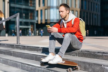 Kırmızı ceketli, beyaz spor ayakkabılı ve yeşil çantalı, modaya uygun sakallı adam portresi. Uzun panolu bir hippi merdivenlerde oturuyor ve cep telefonuyla sohbet ediyor. Seçici odaklanma. Şehir ve sokak fotoğrafı.