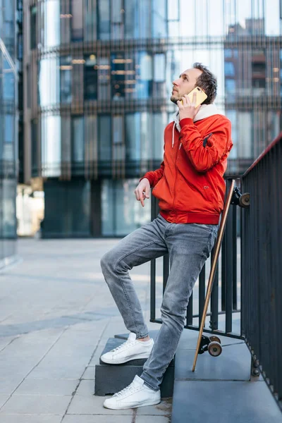 Fashionable man in red jacket, jeans and white sneakers talking on the mobile phone. Longboard is propped up next to it. Selective focus, blurred background. Urban and street photo. Concept of leisure