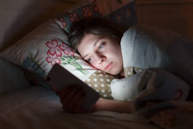 Genç uykulu yorgun kız gece geç saatlerde akıllı telefon kullanarak yatakta uzanıyor, uyuyamıyor. Sosyal medya bağımlılığı, cep telefonu bağımlılığı, uykusuzluk, nomofobi, uyku bozukluğu, uykusuzluk konsepti.