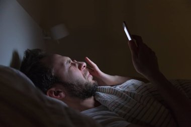 Yetişkin uykucu adam gece geç saatte yatakta internette sörf yapıyor, uyuyamıyor / uykulu yorgun, sosyal medya bağımlısı, cep telefonuna bağımlı, yarı kapalı yorgun gözler, uykusuzluk, uykusuzluk konsepti