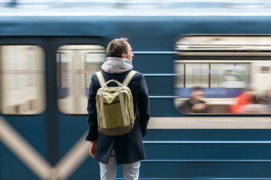 Metro platformunda duran, mavi ceketli, yeşil sırt çantalı, yalnız ve yakışıklı bir adam, hareket halinde. Modern bir şehir sakini için çalışmanın yolu. Sabah işe gidişi. Kentsel yaşam konsepti.