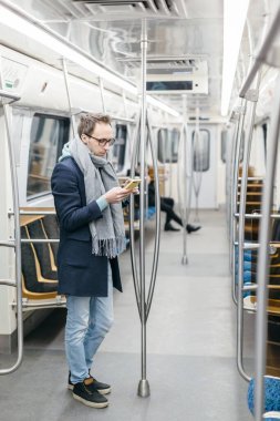Gözlüklü ve kablosuz kulaklıklı bir adamın metro trenine binerken çekilmiş dikey fotoğrafı. O, mavi bir ceket ve gri bir eşarp giyiyor, tırabzan tutuyor ve akıllı telefon kullanıyor. Sosyal ağlar bağımlılığı.