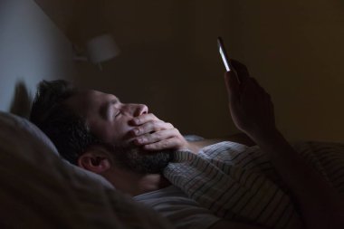 Yetişkin uykulu esneyen erkek gece geç saatte yatakta internette sörf yapıyor, uyuyamıyor / uykulu yorgun, sosyal medya bağımlısı, cep telefonuna bağımlı, yarı kapalı yorgun gözler, uykusuzluk kavramı.