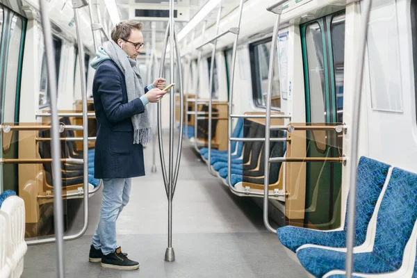 Gözlüklü ve kablosuz kulaklıklı bir erkek metro trenine biniyor. O, mavi bir ceket ve gri bir eşarp giyiyor, tırabzan tutuyor ve akıllı telefon kullanıyor. Dijital bağımlılık, sosyal ağlar bağımlılığı.