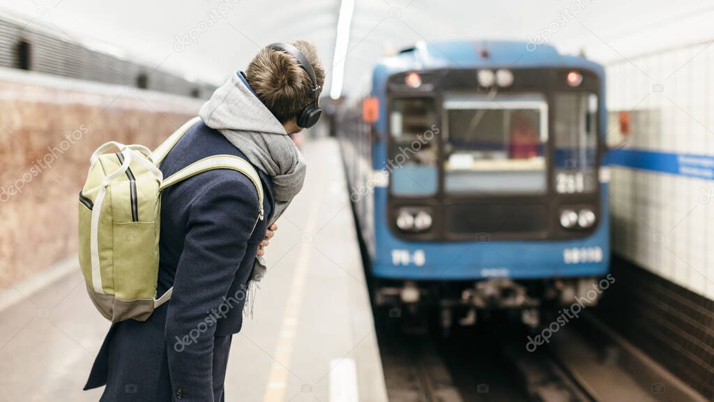 Cropped photo of young handsome man in a blue coat with green backpack behind and headphones on the head looks after the departing subway train. Selective focus on the male.Copy space. Concept of time