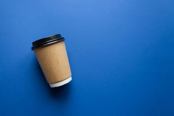 Coffee to go concept. Disposable craft cup with black plastic cover on a trendy classic blue background. Place for text. Flat lay style. Top view.