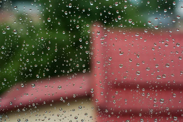 Kapky deště na okenní sklo se stromy a červenou střechou jako pozadí — Stock fotografie