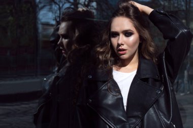 Güneşin batışında şehrin arka planında inanılmaz makyajlı modaya uygun siyah ceket giyen zarif bir kızın güzel portresini kapat. Pencerede yansıma. Metin uzayı.