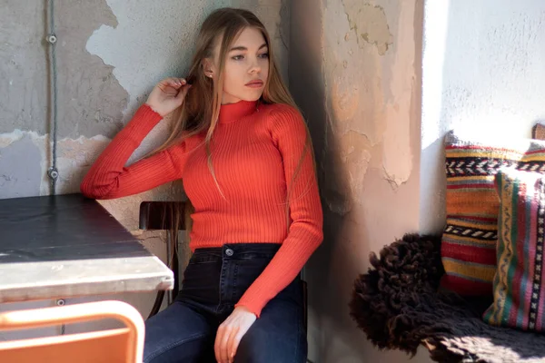 Charming woman with beautiful long hair wearing a orange sweater during rest in coffee shop with an unusual interior. Indoor portrait with light from the window. Text space.