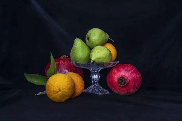 Still life. Pears in a glass vase, oranges and pomegranates on a black background — Stock Photo, Image
