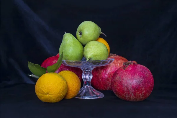 Stillleben. Birnen in einer Glasvase, Orangen und Granatäpfel auf schwarzem Hintergrund — Stockfoto