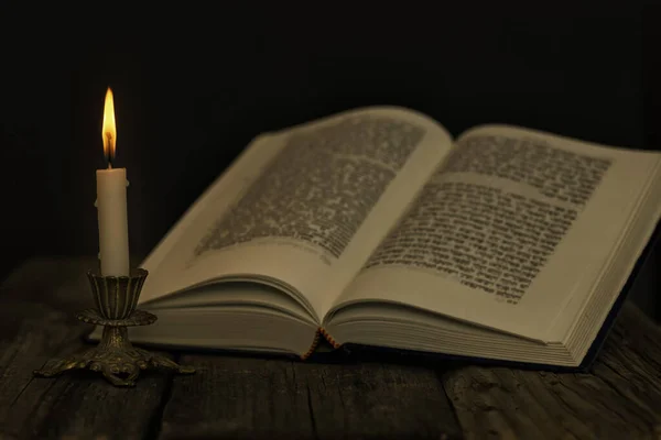 Torá abierta y una vela encendida en un candelero sobre una superficie de madera sobre un fondo oscuro — Foto de Stock
