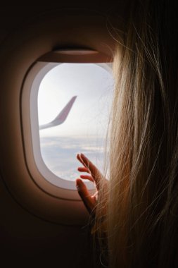 Kadın uçağın penceresinden dışarı bakıyor. Elini pencerenin yanına koy. Uçaktaki kız. Uçak kanadı, manzara manzarası. Güzel bulut, mavi gökyüzü. Uçakla seyahat, macera. Hava ulaşımı. Havacılık