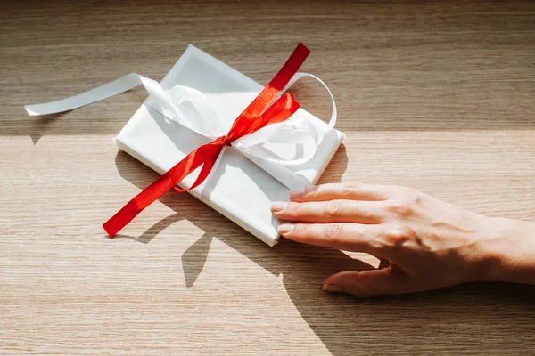 Woman hold gift in hand. Holiday present. Box with surprise. Happy celebration event. Kraft paper packaging. Red bow. Wrapping paper and ribbon. Female hand with box close-up. Handmade congratulation