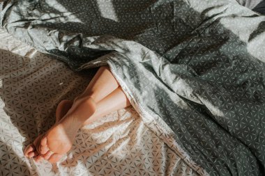 Kadının ayakları yorganın altından dışarı bakıyor. Kız tek başına yatakta uyuyor. Uyan! Uyan! Dişi bacaklar. Yatak odasında mutlu sabahlar. Rahat ve rahat. Yatak çarşaflarında güneş ışığı. Yastık, battaniye. İyileşme, rahatla.