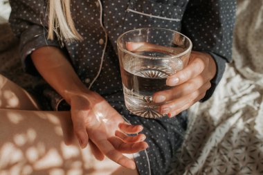 Yatakta elinde bir bardak su tutan bir kadın. İyi sabahlar. Pijamalı kız. Sağlıklı yaşam tarzı, sağlık. Düzgün beslenme. İçme suyu. Suyla sabah. Çarşaflarda güneş ışığı. Yastık, battaniye
