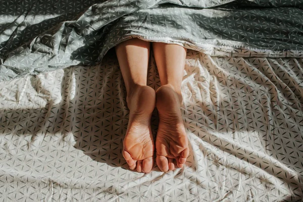 一个女人的脚从被子里探出头来 只有女孩一个人睡在床上 醒醒吧 女性腿 早上好 在卧室里 舒服舒适 床单上的阳光 — 图库照片