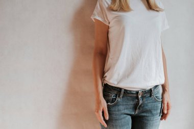 Blond hair woman posing near a light wall. Beautiful young caucasian girl. Hand gestures. Emotion. Casual clothing. Studio model in work. Strong woman, future is female. Jeans and white blank t-shirt