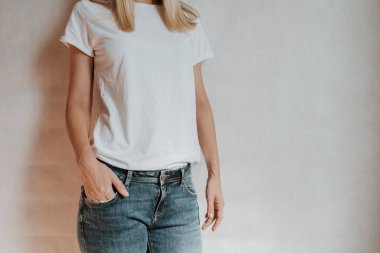 Blond hair woman posing near a light wall. Beautiful young caucasian girl. Hand gestures. Emotion. Casual clothing. Studio model in work. Strong woman, future is female. Jeans and white blank t-shirt
