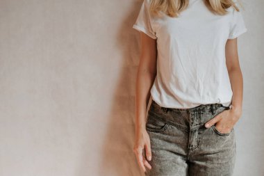 Blond hair woman posing near a light wall. Beautiful young caucasian girl. Hand gestures. Emotion. Casual clothing. Studio model in work. Strong woman, future is female. Jeans and white blank t-shirt