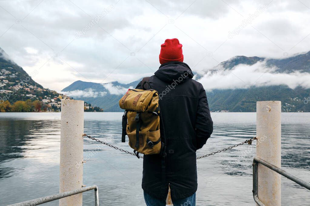 Man with backpack enjoy panorama on pier in Lugano. Man in travel. Lake Lugano, southern slope of Alps. Landscape in Switzerland. Amazing scenic outdoors view. Canton of Ticino. Adventure lifestyle