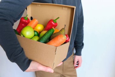 Adamın elinde doğal taze sebzeler ve meyvelerle dolu bir kutu var. Yemek dağıtım servisi. Biyolojik organik besinlerle sağlıklı beslenme ve beslenme. Marketten alışveriş. Vejetaryen yaşam tarzı. Yemek pişirmeye hazır.