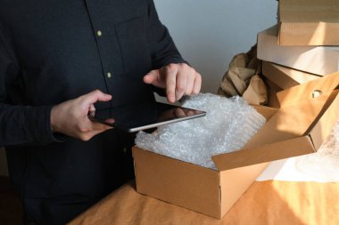 Bir adam karton kutular paketliyor. Online siparişler için teslimat servisi. Tableti elimde tutuyorum. Ofiste çalışan adam, kargo işi. Ürünlü depo, ekspres nakliye hazırlığı