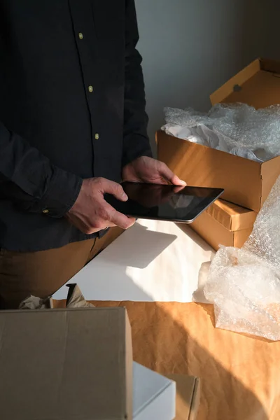 A man is packing cardboard boxes. Delivery service for online orders. Holding tablet in hand. The guy working in office, the cargo business. Warehouse with products, preparation for express shipping