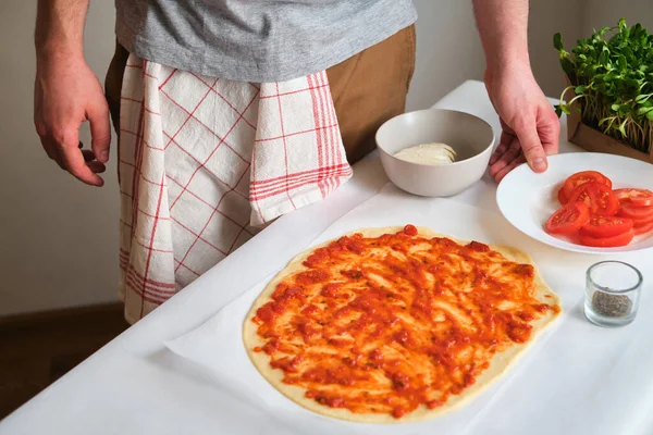 A man is preparing pizza. Cooking homemade italian pizza. Preparation raw ingredients for baking. Sauce, tomatoes and cheese. Fresh natural healthy food. ulinary hef kneading dough on kitchen table
