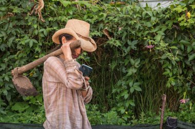 Omzunda çapa tutan şapkalı kadın meyve bahçesinde akıllı telefonuna bakıyor.
