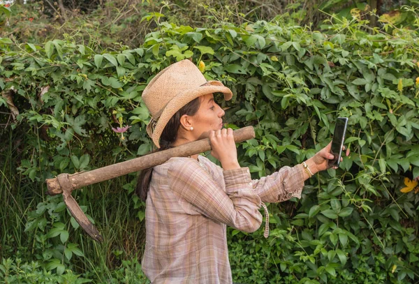 Şapkalı bir kadın omzunda çapayla meyve bahçesinde akıllı telefonuyla selfie çekiyor.