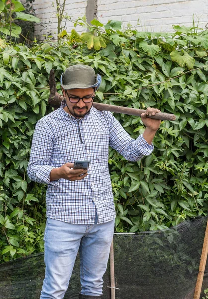 Omzunda çapa tutan şapkalı bir adam meyve bahçesinde akıllı telefonuna bakıyor.
