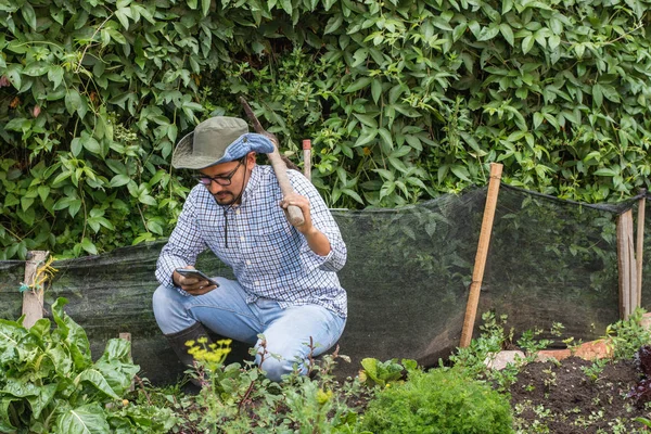 Man crouching with hat holding a hoe over his shoulder looking his smartphone in a orchard