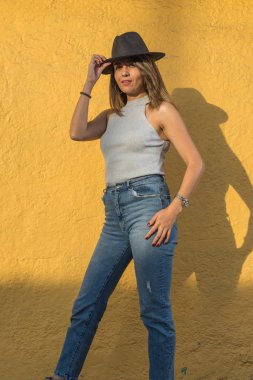 Genç ve güzel bir Latin kadının portresi. Sağ eli sarı duvarda siyah şapkasını alıyor.