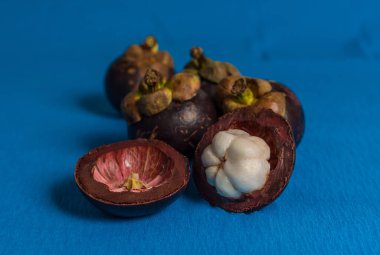 Mangosteen ve enine kesitleri mavi arkaplanda kalın mor deri ve beyaz eti gösteriyor.
