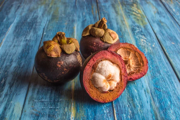 Mangosteen, iki tam meyve ve enine kesit. Eski mavi ahşap masanın üzerindeki kalın mor deri ve beyaz eti gösteriyor.