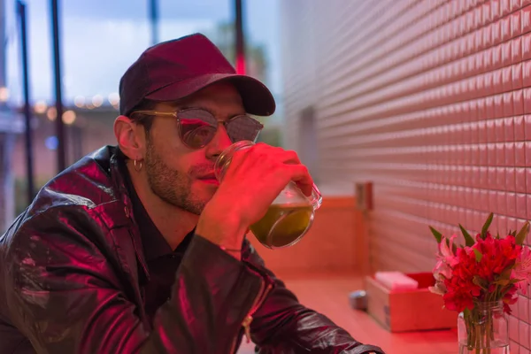 Young attractive latin man with cap, black leather jacket and sun glasses drinking a green healthy detox smoothie from a glass jar in a restaurant with neon lights