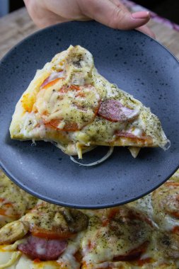 The hand holds a plate with a piece of pizza over the pizza that stands on the wooden board
