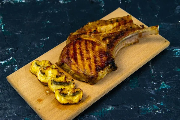 A piece of grilled pork steak on the bone which stands on a wooden board next to stand mushrooms — 스톡 사진