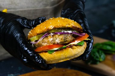 Siyah kauçuk eldivenli bir kadının ellerinde et pirzolası, marul, domates ve doğranmış peynirli sulu bir burger var..