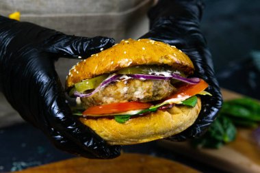 Siyah kauçuk eldivenli bir kadının ellerinde et pirzolası, marul, domates ve doğranmış peynirli sulu bir burger var..