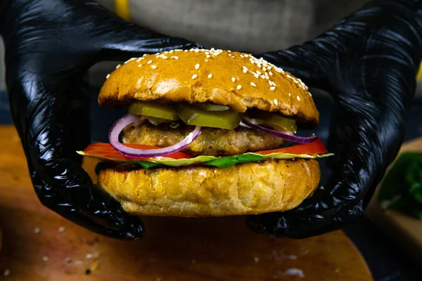 Las manos de mujer con guantes de goma negra sostienen una jugosa hamburguesa con chuleta de carne, lechuga, tomate, queso rallado . — Foto de Stock