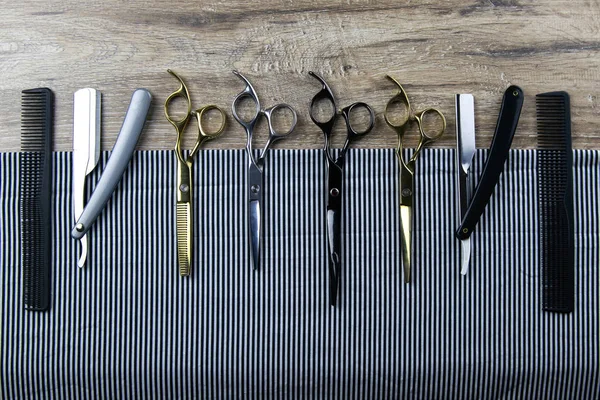 Un conjunto de tijeras para cortes de pelo, peines y una peligrosa cuchilla extendida sobre una toalla a rayas que se apoya sobre una mesa de madera —  Fotos de Stock