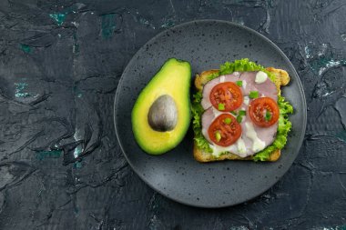 Jambonlu sandviç ve avokadolu gri tabakta domates.