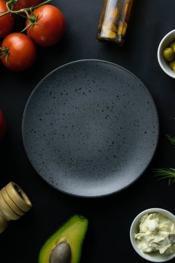 Siyah arka planda gri bir tabak, domates, avokado, zeytin, beyaz sos, yağ ve baharatlar.