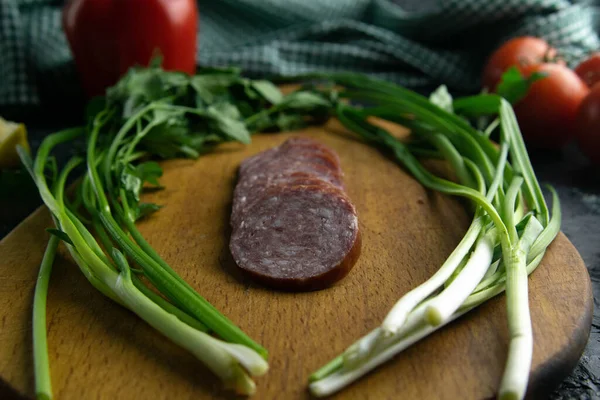 Salami rebanado en una tabla de madera, alrededor del jamón, cebollas verdes, eneldo y perejil . — Foto de Stock